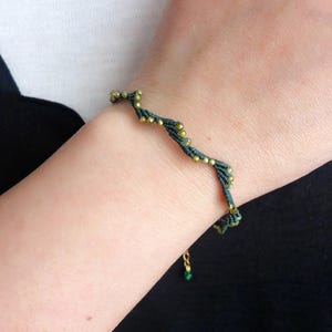以下が含まれることがあります： 暗い緑色のブレスレットに金色のビーズが織り込まれたデザイン。ブレスレットは人の手首につけています。
