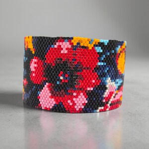 Peut inclure: Un large bracelet de perles avec un motif floral. Le bracelet présente un fond noir avec une grande fleur rouge et des accents de rose, de jaune et de bleu. Le bracelet est fait de petites perles colorées.