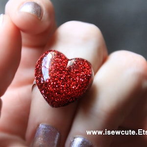 May include: A red glitter heart-shaped ring on a finger. The ring is made of resin and has a shiny finish.