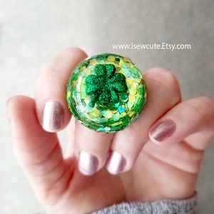 May include: A green four-leaf clover charm encased in a clear resin ring with green and gold glitter. The ring is on a finger with pink nail polish.
