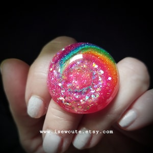 May include: A round resin ring featuring a vibrant pink base filled with iridescent glitter and a swirling rainbow arc of red, orange, yellow, green, and blue colors across the top surface. The text www.isewcute.etsy.com is printed below.