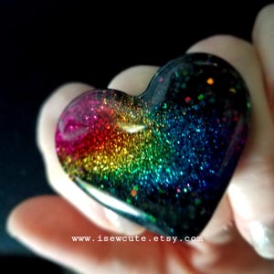 May include: A heart-shaped resin pendant with a rainbow glitter design. The colors transition from pink to red, yellow, green, blue, and purple, set against a black background. The website address www.isewcute.etsy.com is visible at the bottom.