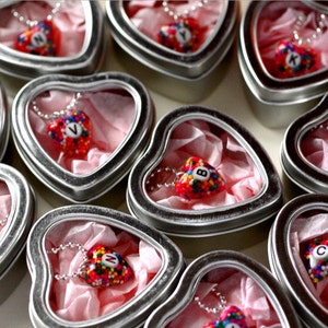 May include: Heart-shaped necklaces with letters A through Z, each encased in a silver heart-shaped tin. The necklaces are red with colorful sprinkles and a silver chain. The tins are lined with pink tissue paper.