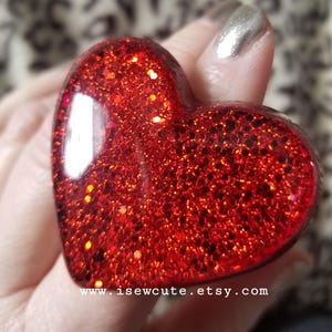 May include: A red heart-shaped ring with red glitter embedded in the resin. The ring is on a finger.