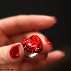 May include: A heart-shaped ring with a red glitter design. The ring is held in a hand with red nail polish. The ring is a romantic accessory.