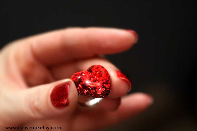 May include: A heart-shaped ring with a red glitter design. The ring is held in a hand with red nail polish. The ring is a romantic accessory.