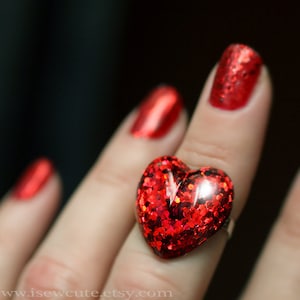 May include: A close-up of a red heart-shaped ring with glitter. The ring is on a finger with red nail polish. The background is dark. The ring is a statement piece.