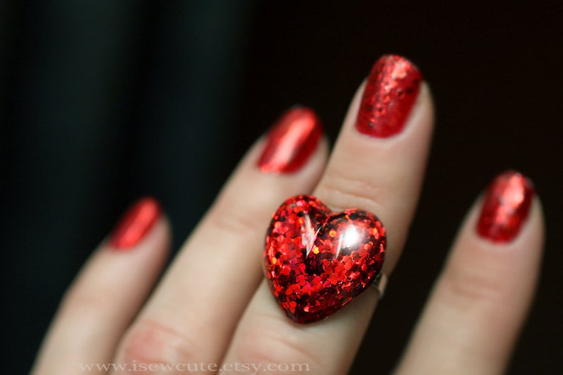 May include: A close-up of a red heart-shaped ring with glitter. The ring is on a finger with red nail polish. The background is dark. The ring is a statement piece.