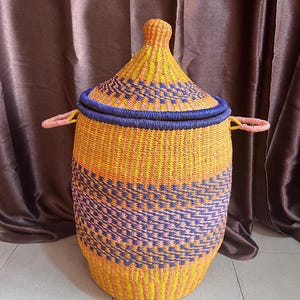 May include: A woven storage basket with a lid. The basket is primarily yellow and orange, with blue and pink geometric patterns. It has two pink handles and a blue rim around the lid. The basket is made of natural fibres.