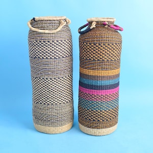 Op de afbeelding: Twee hoge, geweven manden met handvatten. Eén is voornamelijk zwart en bruin met een ruitpatroon. De andere heeft horizontale strepen in geel, blauw, turquoise en roze. Beide hebben een cilindrische vorm en staan tegen een lichtblauwe achtergrond.