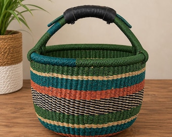 Puede incluir: Una cesta redonda tejida con rayas horizontales en tonos verde bosque, azul cerceta, naranja quemado y beige natural. La cesta tiene un asa arqueada única envuelta en cuero negro en el centro, lo que proporciona un agarre firme al llevarla.