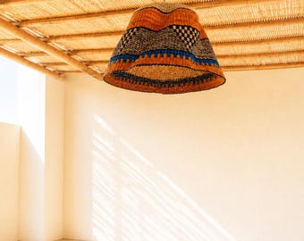 Pode incluir: Um abajur suspenso com textura trançada e silhueta em forma de sino. O exterior apresenta faixas horizontais nas cores laranja, preto e azul, com um padrão geométrico xadrez integrado na seção superior.