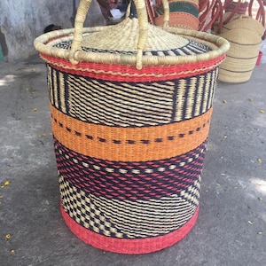 May include: A cylindrical woven basket with a lid and handle. The basket features bands of orange, black, red, and cream colours. The lid and handle are made of natural-coloured woven material.