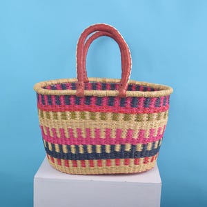 May include: A woven basket with two leather-wrapped handles featuring a horizontal striped pattern in shades of pink, navy blue, olive green, and natural tan. The basket has an oval opening and a structured base, crafted from natural fibers.