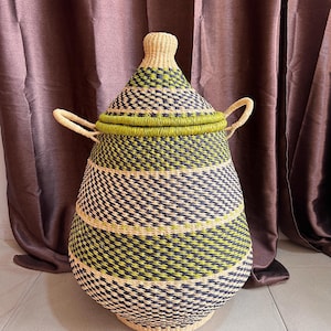 May include: A woven storage basket with a lid. The basket is a light tan colour with a pattern of black and green woven stripes. The lid has a handle and is also woven. The basket has two handles on the sides.