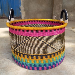 May include: A large, colorful woven basket with two black and white handles. The basket is made of natural materials and has a pink, yellow, black, blue, and green pattern.