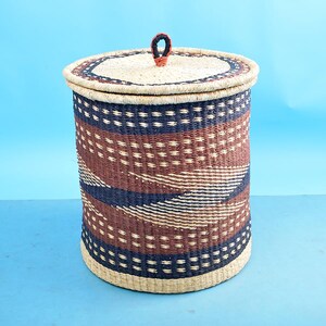May include: A woven storage basket with a lid and a handle. The basket is made of natural materials and has a brown, blue and white geometric pattern.