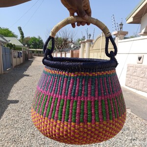 May include: A large, woven basket with a pink, green and yellow striped pattern. The basket has a black handle and is made of natural materials.