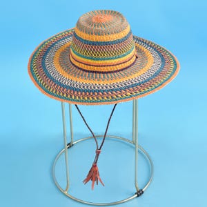以下が含まれることがあります： オレンジ、黄色、青、白のストライプが特徴の、幅広のつばが付いたカラフルな織りのサンハット。帽子にはタッセル付きの茶色のあご紐が付いており、金色の金属製のスタンドに飾られています。夏のアクセサリー。