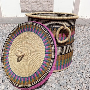 Può includere: Un cesto di vimini con coperchio per la conservazione. Il cesto è realizzato con materiali naturali e presenta un motivo a strisce rosa, marrone e verde. Il coperchio ha un motivo simile e una maniglia intrecciata.