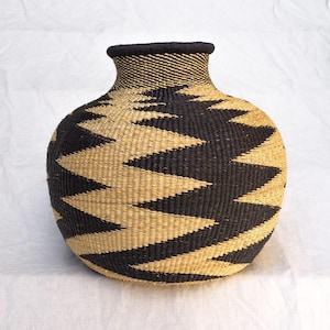 May include: A round, hand-woven basket vase featuring a wide, bulbous body and a narrow neck. The exterior displays a repeating, high-contrast zigzag pattern in black and natural tan fibers, with a solid black rim encircling the top opening.
