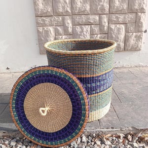 May include: A woven storage basket with a lid. The basket is made of natural materials and has a blue, green, and brown striped pattern. The lid is round and has a similar pattern.