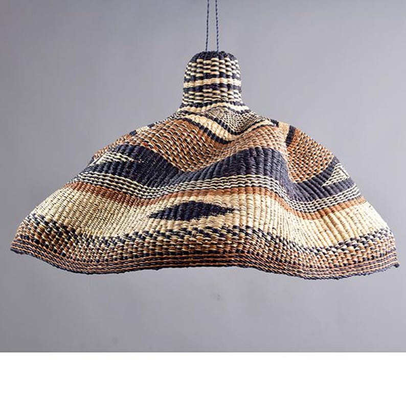 Pode incluir: Uma lumin&aacute;ria pendente tecida com uma sombra larga e flare. A sombra &eacute; feita de fibras naturais em tons de bege, marrom e azul marinho, criando um padr&atilde;o listrado. A lumin&aacute;ria &eacute; suspensa por um cabo escuro e tem um topo cil&iacute;ndrico.