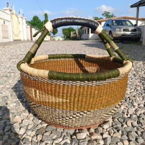 Puede incluir: Una cesta tejida con un dise&ntilde;o marr&oacute;n, verde y negro. La cesta tiene un asa de cuero negro y est&aacute; sentada sobre un lecho de peque&ntilde;as piedras.