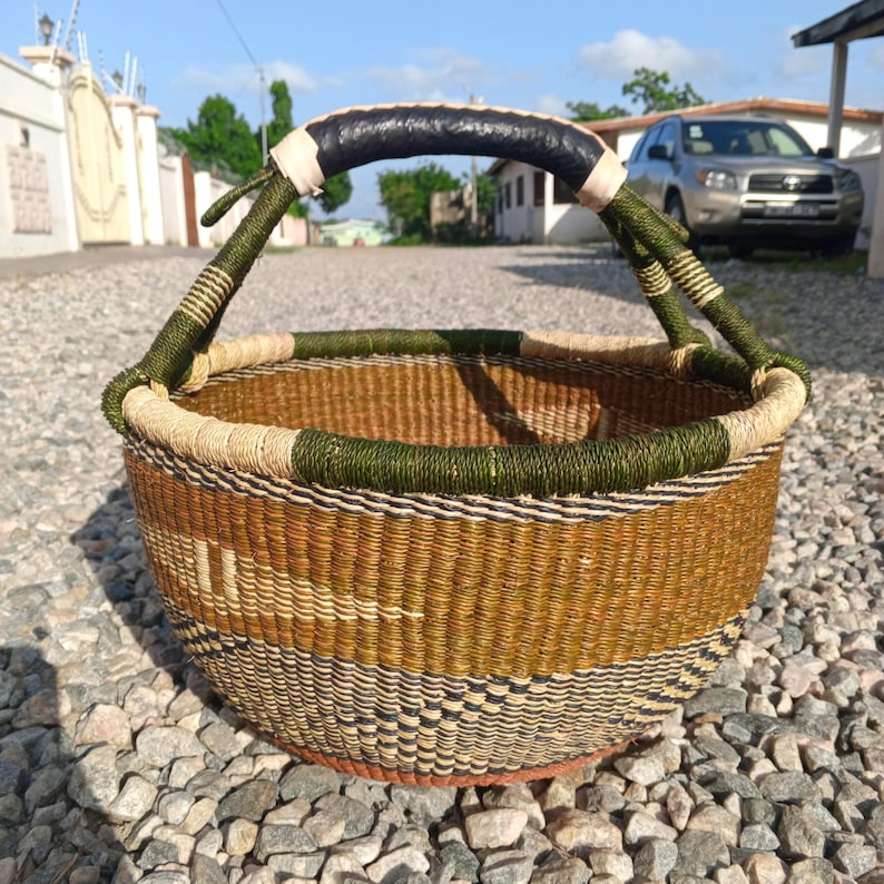 Puede incluir: Una cesta tejida con un dise&ntilde;o marr&oacute;n, verde y negro. La cesta tiene un asa de cuero negro y est&aacute; sentada sobre un lecho de peque&ntilde;as piedras.