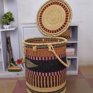 May include: A large woven storage basket with a lid. The basket is made of natural materials and has a black, orange, and pink geometric pattern. The lid is round and has a black and orange striped pattern.