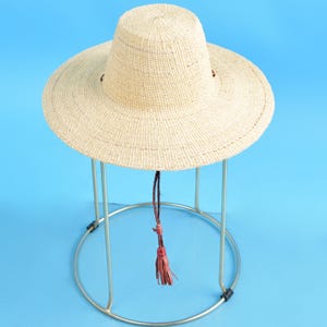 Pode incluir: Um chapéu de palha de aba larga com uma borla de couro marrom. O chapéu é de cor marrom claro e tem uma textura tecida. É exibido em um suporte de metal prateado.