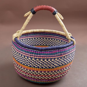 Può includere: Un grande cesto rotondo intrecciato con un motivo a strisce colorate in tonalità di blu, rosa, verde, arancione e giallo. Il cesto ha una robusta maniglia in pelle.