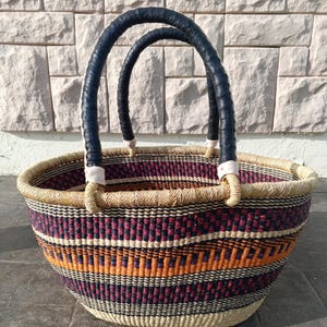 May include: A woven oval basket with black leather handles. The basket has a colourful striped pattern in shades of orange, purple, and white.