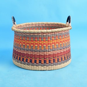 May include: A round woven basket with a colorful geometric pattern in red, orange, yellow, and navy blue. The basket has two dark blue handles and a natural fiber rim. The basket is approximately 14 inches in diameter and 12 inches tall.