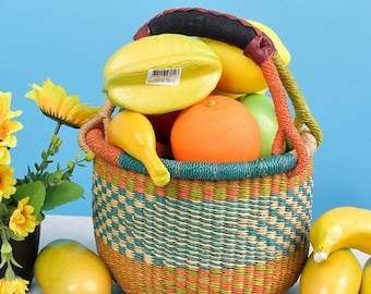 Puede incluir: Una cesta tejida colorida llena de carambolas amarillas, plátanos amarillos, una manzana verde y una naranja brillante. La cesta tiene un patrón azul, verde, naranja y rojo.