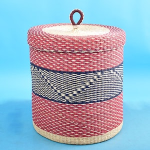 May include: A red and blue woven storage basket with a lid and a leather handle. The basket is made of natural materials and has a geometric pattern.