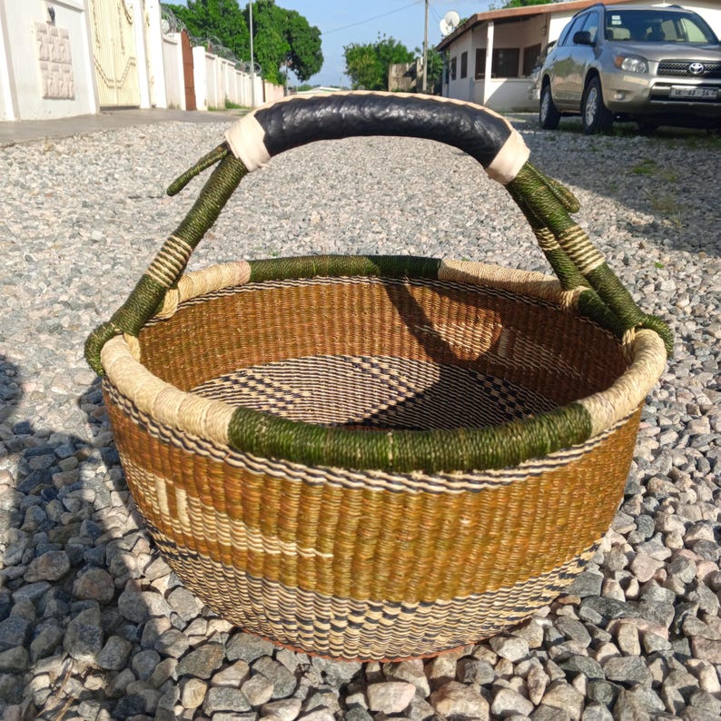 Puede incluir: Una cesta grande, redonda y tejida con un asa de cuero negro. La cesta est&aacute; hecha de materiales naturales y tiene una combinaci&oacute;n de colores marr&oacute;n, verde y negro.