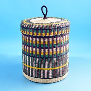 May include: A woven storage basket with a lid and a handle. The basket is made of natural materials and has a colourful geometric pattern in shades of green, brown, and yellow.