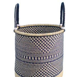 May include: A cylindrical woven basket with a natural beige base and rim. The body features a repeating geometric pattern in navy blue and beige. Two navy blue handles with white accents rise from the rim, offering a stylish storage solution.