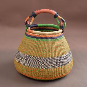 May include: A handwoven basket with a rounded base and a handle. The basket features a mix of colors, including orange, green, blue, and tan. The handle is wrapped in brown leather. The basket is likely used for storage or decoration.