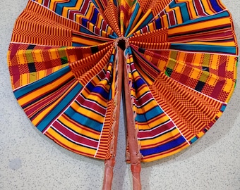 Könnte beinhalten: Ein runder Fächer mit einem lebendigen Kente-Stoffmuster aus geometrischen Streifen und Blöcken in Gelb, Orange, Rot, Blau und Grün. Der Fächer besteht aus plissiertem Stoff, der an zwei braunen Ledergriffen befestigt ist, die durch einen Lederriemen verbunden sind.