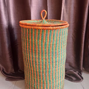 May include: A tall, cylindrical woven basket with a lid. The basket is made of orange and green striped material. The lid has a loop handle. The basket is likely used for storage or laundry.