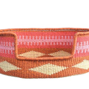 May include: Woven pet bed in shades of orange, pink, and cream. The bed features a low front and a raised back, with a diamond pattern on the base. The sides have a geometric design.