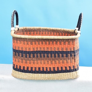 Puede incluir: Una cesta de almacenamiento tejida de color naranja, negro y marrón con dos asas de cuero negro. La cesta tiene forma ovalada y tiene una base de paja natural.