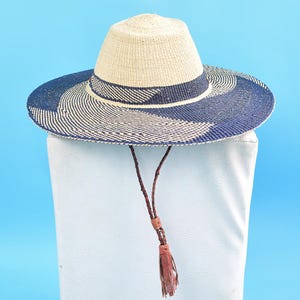 May include: A wide-brimmed straw hat with a blue and white woven pattern. The hat has a brown leather chin strap with a tassel.