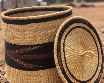 May include: A woven storage basket with a lid. The basket is cylindrical with a black band and decorative stitching. The lid is round with a handle. The basket is made of natural fibers and is sitting on a bed of rocks.