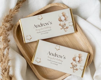 May include: Two rectangular chocolate bars with gold foil edges are displayed on a wooden tray. The bars have a cream-colored background with text and a teddy bear illustration holding balloons. The text reads "Andrew's beary first birthday".