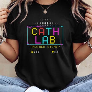May include: A black short-sleeve t-shirt featuring a colorful pixelated graphic that reads CATH LAB, followed by the text ANOTHER STEMI? and two options with icons: a thumbs up labeled Yes and a thumbs down labeled No, all in a retro style.