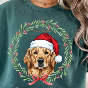 Peut inclure: Un sweat-shirt vert d'eau avec un Golden Retriever portant un bonnet de Père Noël, entouré d'une couronne de Noël de feuilles vertes et de baies rouges. Les mots "Merry Christmas" sont écrits au-dessus du chien, avec un nœud rouge en bas.
