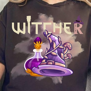 Może przedstawiać: Ciemnoszara koszulka z napisem "WITCHER" w stylizowanej czcionce. Projekt zawiera fioletowy kapelusz czarownicy z grzybami, butelkę z eliksirem i małą ikonę kapelusza czarownicy. Ogólnym motywem jest fantasy i magia.
